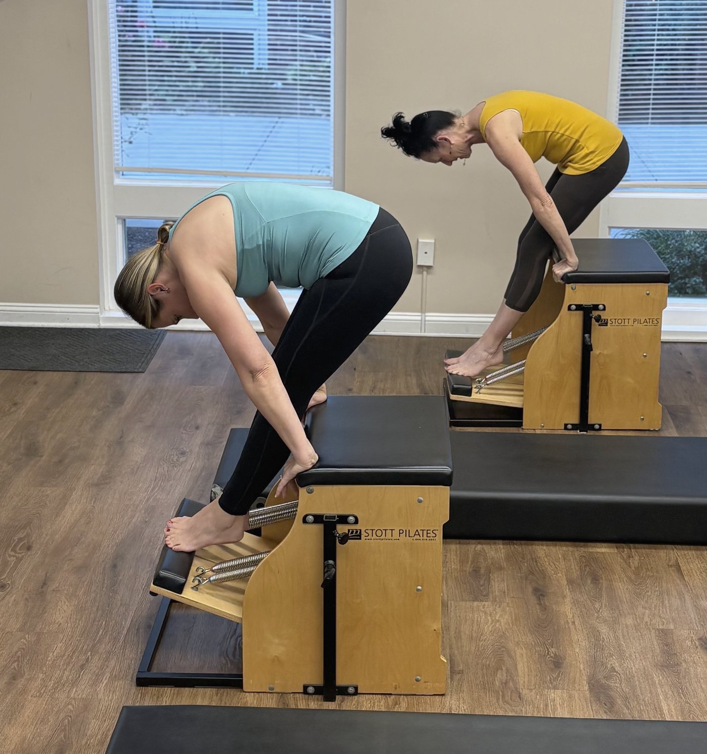 Nicole and a client bending forward on wooden Pilates chairs.